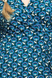 Close-up of the patterned fabric on the BUTIK KUBIK Cissi och Selma dress Vera Miniblom blåbär, featuring blue and teal flowers with white centers and stems against a dark green background. The image shows part of this ekologisk bomullsklänning with a crossover (omlottklänning) neckline, along with a section of light brown hair.