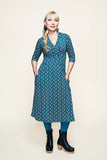 A woman with blonde hair in a braid wears the Cissi och Selma dress Vera Miniblom blåbär by BUTIK KUBIK, styled with teal tights, black platform boots, and green earrings. She stands against a plain light background with her hands in the dress pockets, smiling slightly.