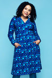 A woman wears a Cissi och Selma dress Viveca Droppe blå, a long-sleeved, knee-length 70s-style shirt dress in blue with a repeating pattern of teardrop shapes and dots, a button-front collar, and two front patch pockets.