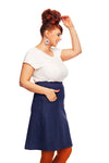 Smiling woman with red hair in a bun, wearing a white top and the Cissi och Selma Majken skirt Jeans from BUTIK KUBIK, orange tights, and flower-shaped earrings, stands against a white background with one hand touching her hair.
