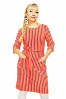 A person with long braided hair stands confidently, wearing a Cissi och Selma dress Doris Ranka by BUTIK KUBIK, featuring red and white stripes and pockets. They sport a cheerful expression with hoop earrings against a plain white background, embodying Cissi och Selmas vårkollektion 2025.