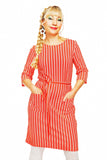 A person with long braided hair stands confidently, wearing a Cissi och Selma dress Doris Ranka by BUTIK KUBIK, featuring red and white stripes and pockets. They sport a cheerful expression with hoop earrings against a plain white background, embodying Cissi och Selmas vårkollektion 2025.