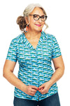 A woman with long gray hair, wearing glasses and red earrings, smiles while buttoning a Cissi och Selma Margit blus Havsbris by BUTIK KUBIK, made from eco-friendly cotton with a sailboat pattern. She pairs it perfectly with blue jeans.