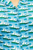 The Cissi och Selma Margit blus by BUTIK KUBIK features a kortärmad skjortblus with blue and green nautical patterns of sailboats and waves. Made from ekologisk bomull, it includes blue buttons, a visible collar, and a light summery design inspired by 50-talets grafiska design.