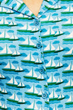 The Cissi och Selma Margit blus by BUTIK KUBIK features a kortärmad skjortblus with blue and green nautical patterns of sailboats and waves. Made from ekologisk bomull, it includes blue buttons, a visible collar, and a light summery design inspired by 50-talets grafiska design.