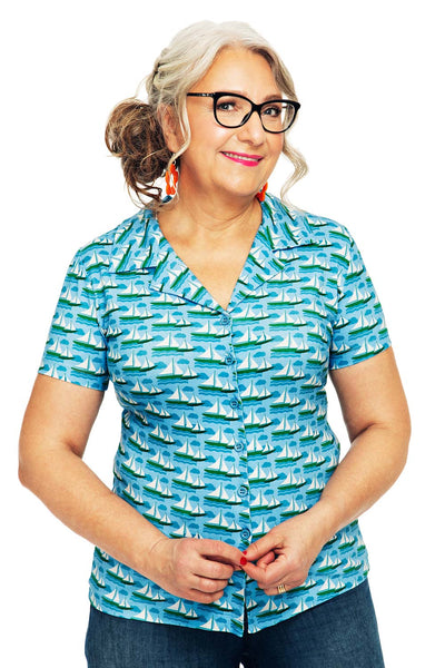 A woman with long gray hair, wearing glasses and red earrings, smiles while buttoning a Cissi och Selma Margit blus Havsbris by BUTIK KUBIK, made from eco-friendly cotton with a sailboat pattern. She pairs it perfectly with blue jeans.