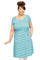 A smiling brown-haired woman with an updo wears a BUTIK KUBIK Cissi och Selma Asta Havsbris dress, featuring boats on blue ekologisk bomullstrikå. With one hand extended, she dons white tights and green earrings against a white backdrop.