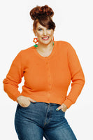 A smiling woman with a red beehive hairstyle wears the Cissi och Selma new cardigan Apelsin by BUTIK KUBIK, paired with blue jeans and orange flower earrings. She stands against a white background, hands on hips.