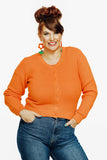 A smiling woman with a red beehive hairstyle wears the Cissi och Selma new cardigan Apelsin by BUTIK KUBIK, paired with blue jeans and orange flower earrings. She stands against a white background, hands on hips.