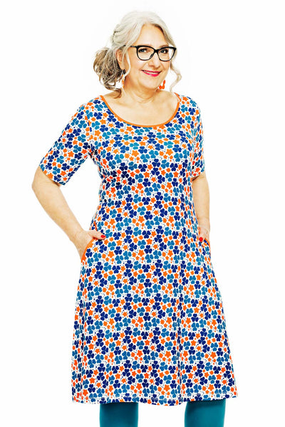 A gray-haired individual with glasses smiles in the Cissi och Selma Magda dress Klöver Orange from BUTIK KUBIK, displaying a multicolored floral design of orange, blue, and red inspired by 50-talets grafiska design. The short-sleeved dress is made from ekologisk bomullstrikå, featuring practical pockets on a white backdrop.