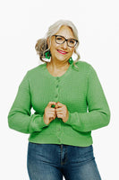 An older woman with long gray hair, glasses, and green earrings smiles while clasping her hands. Shes wearing the Cissi och Selma cardigan Sigrid Björk by BUTIK KUBIK and blue jeans, standing against a plain white background.