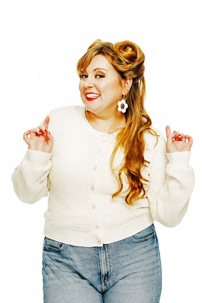 A woman with long, wavy hair smiles and points upwards, wearing a Cissi och Selma cardigan Sigrid Vide from BUTIK KUBIK and jeans. Her large, flower-shaped earrings add playfulness to her look against a plain white backdrop.