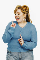 A woman with styled hair and red lipstick smiles playfully. Shes wearing the Cissi och Selma cardigan Sigrid Himmel from BUTIK KUBIK, large white flower earrings, and denim jeans against a plain white background.