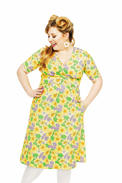 A woman confidently poses against a white background, wearing a vibrant floral Cissi och Selma Vivi dress humlesurr gul from BUTIK KUBIK. Her vintage curls and large white drop earrings complement the knee-length skirt as she smiles radiantly with her hands on her hips.