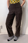 An individual stands with hands in the pockets of brown high-waisted Kubik corduroy trousers by BUTIK KUBIK. Theyre wearing a retro-patterned shirt and black-and-white checkered socks, set against a simple, light-colored wall and floor background.