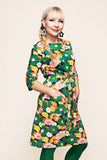 A smiling woman with a blonde braid models the Cissi och Selma dress Doris 1970 Grönorange from BUTIK KUBIK—an A-line, 60s-inspired green floral dress adorned with pink, orange, and yellow flowers, made from organic cotton jersey. She completes the look with large blue earrings and green tights, posing with her hands in the dress pockets against a cream background.