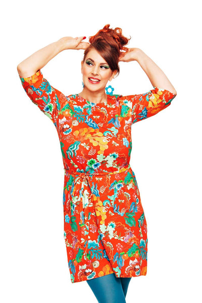 A red-haired individual with a bun poses playfully in the vibrant Cissi och Selma dress Doris Evighet Orange by BUTIK KUBIK and blue tights. They smile, gaze off to the side, and frame their face with both hands against a plain white background.