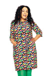 A woman with curly hair smiles and poses with one hand on her hip, wearing the Cissi och Selma dress Doris Fågelskvaller from BUTIK KUBIK and green tights against a white background.