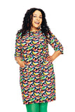 A woman with curly hair smiles and poses with one hand on her hip, wearing the Cissi och Selma dress Doris Fågelskvaller from BUTIK KUBIK and green tights against a white background.