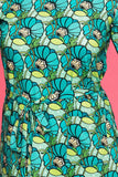 A person wears the Cissi och Selma dress Doris Kronblad Turkos from BUTIK KUBIK, a bold teal and green floral A-line dress in organic cotton with a waist tie and large stylized flowers, standing against a pink background.