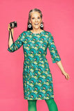 A smiling woman with gray hair, in a Cissi och Selma dress Doris Kronblad Turkos from BUTIK KUBIK, green tights, and white flower earrings, holds a vintage camera against a bright pink background.