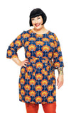 A smiling woman with short black hair wears the Cissi och Selma dress Doris Retroblom Blåbrun from BUTIK KUBIK, orange tights, and displays forearm tattoos. She stands against a white background with one hand on her hip.