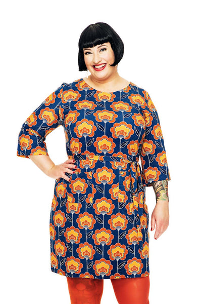 A smiling woman with short black hair wears the Cissi och Selma dress Doris Retroblom Blåbrun from BUTIK KUBIK, orange tights, and displays forearm tattoos. She stands against a white background with one hand on her hip.