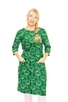 A blonde woman with a side braid, smiling slightly and looking up thoughtfully, stands with her hands in her pockets wearing the Cissi och Selma dress Doris Retroblom Grön by BUTIK KUBIK. The background is plain white.
