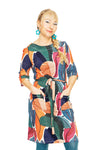 A woman smiles with hands in her pockets, wearing the Cissi och Selma dress Doris Skogsdröm from BUTIK KUBIK—a colorful, 60s-inspired A-line cotton dress with a belt. She pairs it with teal earrings, tights, and a side braid against a white background.
