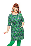 A cheerful redhead poses against a white backdrop in a 60-talet-inspired Cissi och Selma dress Doris Vår Gata Grön by BUTIK KUBIK. Her vibrant, animated expression complements large green earrings and tights.