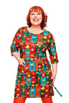 A person with short curly red hair smiles in a colorful Cissi och Selma dress Doris Växtkraft brun, inspired by the 60s and crafted from ekologisk bomullstrikå with handy pockets. The background is plain white. Brand: BUTIK KUBIK.