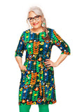 Smiling woman with long gray braided hair, wearing glasses and a colorful Cissi och Selma dress Doris Växtkraft grön from BUTIK KUBIK, stands with one hand on her hip against a white background.
