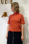 A person with short, wavy hair and glasses wears the BUTIK KUBIK Kubik t-shirt striped in red and orange, paired with a black skirt with front pockets, standing by a white wall with wooden decor and looking to the left.