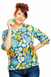 A smiling red-haired woman wears the Cissi och Selma Elsie top Blomklasar by BUTIK KUBIK, a colorful floral A-line top with 3/4 sleeves in blue, orange, yellow, and white, paired with blue jeans and holding a green flower-shaped earring.