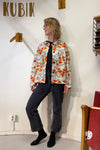 A person stands indoors by a white wall featuring the word KUBIK, wearing glasses, a black shirt, dark jeans, black socks, and the Kubik blouse vintage fabric from BUTIK KUBIK layered on top.