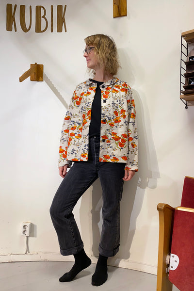 A person stands indoors by a white wall featuring the word KUBIK, wearing glasses, a black shirt, dark jeans, black socks, and the Kubik blouse vintage fabric from BUTIK KUBIK layered on top.