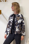 A woman with blonde, wavy hair and glasses wears a black top, black jeans, and the BUTIK KUBIK Kubik Overshirt vintage patterned in black, white, and brown floral deadstock fabric. She stands against an off-white wall, looking to the side.