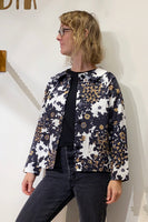 A woman with blonde, wavy hair and glasses wears a black top, black jeans, and the BUTIK KUBIK Kubik Overshirt vintage patterned in black, white, and brown floral deadstock fabric. She stands against an off-white wall, looking to the side.