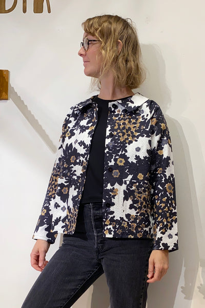 A woman with blonde, wavy hair and glasses wears a black top, black jeans, and the BUTIK KUBIK Kubik Overshirt vintage patterned in black, white, and brown floral deadstock fabric. She stands against an off-white wall, looking to the side.