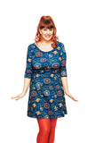 A smiling woman with red hair in loose waves wears the BUTIK KUBIK Cissi och Selma dress Ester Tivoli petrol, made from organic cotton jersey, with orange tights, standing arms outstretched against a white background.