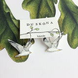 A pair of Du sköna bird earrings Fiskmås by BUTIK KUBIK, featuring a handpainted seagull hoop design on a display card, set against an illustrated leafy background.