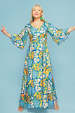 A smiling woman stands barefoot against a light blue background, wearing the Cissi och Selma Greta klänning Blomklasar by BUTIK KUBIK—a long, colorful floral dress with trumpet sleeves and bright yellow, orange, and white flowers.