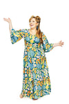 A woman with long, wavy red hair wears the colorful Cissi och Selma Greta klänning Blomklasar from BUTIK KUBIK, standing barefoot and smiling with one hand raised against a plain white background.