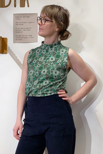 A person wearing glasses stands confidently, sporting the Kubik top cineast spring onion by BUTIK KUBIK and dark trousers. They pose with one hand on their hip, looking to the side. A sign with text is visible in the background.