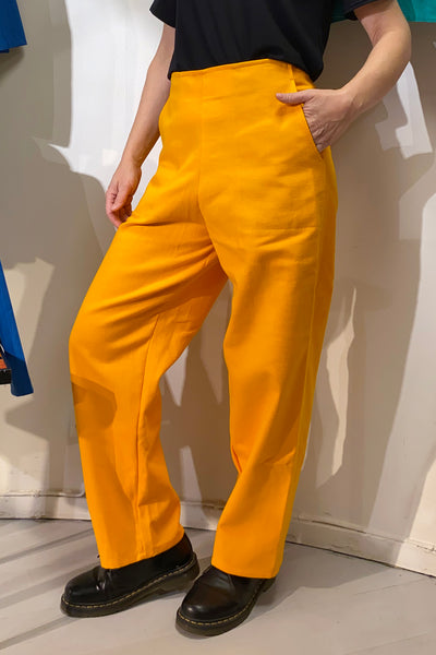 Wearing BUTIK KUBIK’s Kubik trousers yellow vintage, a person pairs the bold high-waisted wide-leg pants with a black top and shoes, standing against a light wall and channeling French workwear inspiration.