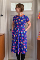 Standing before a white door, a person models the Kubik dress Twist Anemone blue by BUTIK KUBIK. This charming 50s-inspired dress features colorful floral patterns, short sleeves, convenient pockets, and is made from oekotex-certified fabric. Short hair and dark tights complement the look against a glimpse of a wooden cabinet.