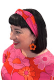 A smiling woman with black hair wears a bright floral top, flower earrings, and the Cissi och Selma Hedda headband 1960 Rosa by BUTIK KUBIK, looking to the left against a white background.