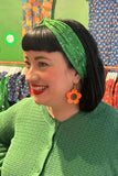 A smiling woman with black hair wears the Cissi & Selma Hedda hårband Ängsblom grön from BUTIK KUBIK, along with bright orange flower-shaped earrings. Colorful patterned dresses hang in the background.