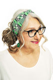 An older woman with gray hair and glasses smiles slightly to the side while wearing a Cissi & Selma Hedda hårband Brygga from BUTIK KUBIK and green earrings. She is dressed in a white top against a plain white background.