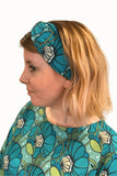 A woman with shoulder-length blonde hair wears the Cissi och Selma Hedda headband Kronblad turkos by BUTIK KUBIK, looking left against a plain white background.
